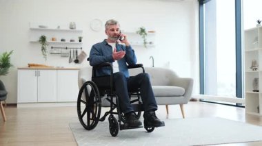 Tekerlekli sandalyede oturan olgun beyaz adam ev ortamında cep telefonuyla konuşuyor. Gündelik yaşam anı parlak ve modern bir iç dekorla sunuluyor..