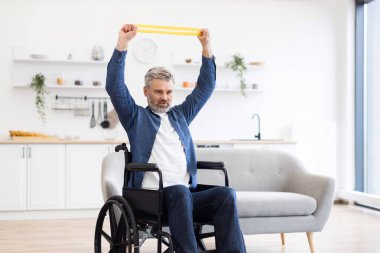 Tekerlekli sandalyede oturan, lastik direnç bandı takan, fitness egzersizi yapan ve modern ve aydınlık bir evde aktif yaşamı teşvik eden olgun bir beyaz adam.