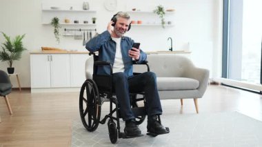 Tekerlekli sandalyedeki olgun beyaz adam, modern yaşam alanında kulaklık ve telefon kullanarak müzik dinlemekten hoşlanıyor. Müzik, bağımsızlık ve teknolojinin kullanımına olan tutkusunu ifade eder.