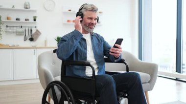 Tekerlekli sandalyedeki olgun beyaz adam telefonu kullanıyor, kulaklıkla müzik dinliyor, gülümsüyor ve kutluyor. İçi rahat modern ev ortamları. Neşe müziği ve erişilebilirlik artışı kavramı.