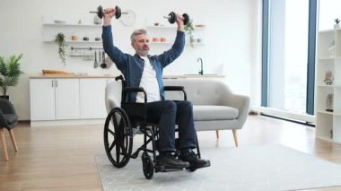 Evde spor yaparken dambıl kaldıran tekerlekli sandalyedeki olgun beyaz adam pozitiflik, güç ve esneklik gösteriyor. Doğal ışıklandırması olan parlak ve modern bir ortam