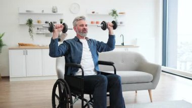 Tekerlekli sandalyede oturan olgun, beyaz bir adam ev içinde dambıllarla egzersiz yapıyor, fitness ve kararlılığı teşvik ediyor..