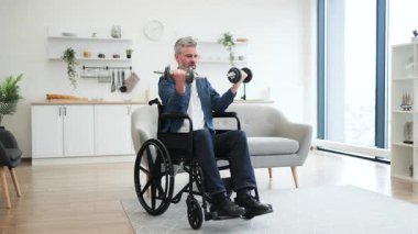 Tekerlekli sandalyede oturan olgun bir beyaz adam içeride halterlerle egzersiz yapıyor. Orta yaşlı bir yetişkin, fiziksel kondisyon ve sağlıklı yaşam tarzını destekliyor.