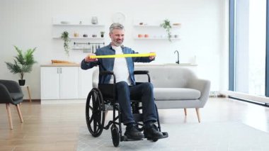 Tekerlekli sandalyede oturan, lastik direniş bandıyla egzersiz yapan, rahat modern oturma odasında esneme egzersizi yapan olgun beyaz bir adam. Sahne fitness ve rehabilitasyonu vurguluyor