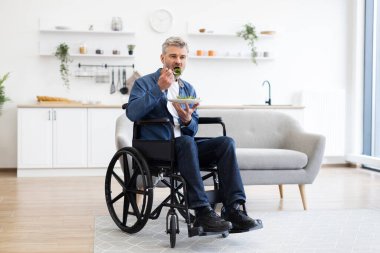 Tekerlekli sandalyede oturan orta yaşlı bir adam parlak bir oturma odasında salatanın tadını çıkarıyor. Sahne kapsayıcılığı, ulaşılabilirliği ve sağlıklı yeme tarzını vurgular. Modern mobilyalı beyaz mutfak.