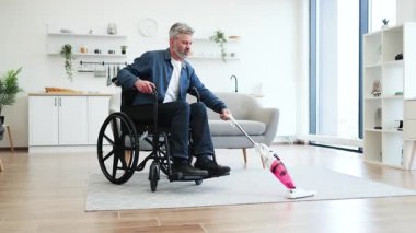 Orta yaşlı, tekerlekli sandalyede beyaz bir adam modern oturma odasında halı süpürüyor, gülümsüyor, takım çalışması yapıyor.