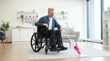 Orta yaşlı, tekerlekli sandalyede temizlik halısı içinde elektrikli süpürgeyle modern ev içinde yaşayan bir adam. Bağımsız yaşam, ulaşılabilirlik, evde bakım ve kararlılığı gösterir.