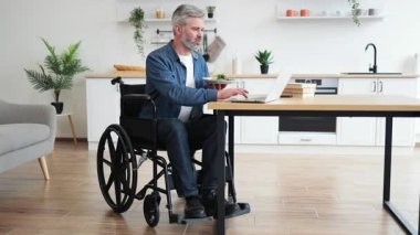 Modern mutfakta dizüstü bilgisayarla çalışırken tekerlekli sandalyede salata yiyen orta yaşlı bir adam. Görüntü, kapalı alanda engelli bireyler için bağımsızlık, üretkenlik ve ev çalışma alanını yansıtır