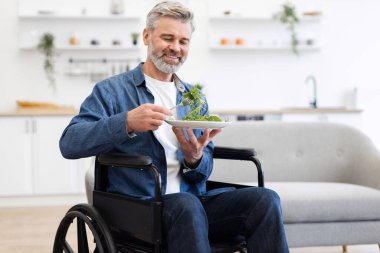 Tekerlekli sandalyede oturan kıdemli adam içeride taze salata yiyor. Sağlıklı yaşam tarzı, ulaşılabilirlik, pozitif yaşlanma, bağımsızlık kavramı. Modern parlak iç plan, olgun ve mutlu birey.