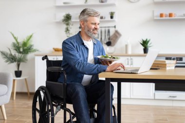 Evde dizüstü bilgisayarla çalışırken tekerlekli sandalyede sağlıklı salata yiyen olgun bir adam. Üretkenliği, bağımsızlığı ve iç çevrede besleyici yeme alışkanlıklarını teşvik eden günlük ortam.
