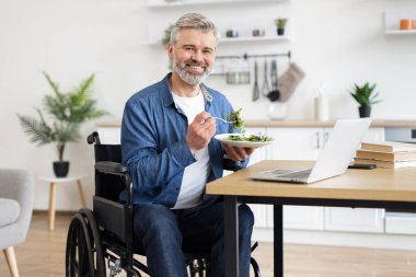 Tekerlekli sandalyedeki yaşlı adam taze salata yiyor, dizüstü bilgisayarda çalışıyor, içeride çalışıyor. Aktif yaşam tarzı, bağımsızlık, sağlıklı yeme alışkanlıkları, ev işi ortamı gösterir.