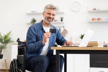 Tekerlekli sandalyede oturan yaşlı adam evdeki online işlemlerde dizüstü bilgisayar ve kart kullanıyor.