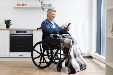 Ekose battaniyeli, tekerlekli sandalyedeki olgun adam akıllı telefonu açık havada, modern mutfakta kullanıyor.