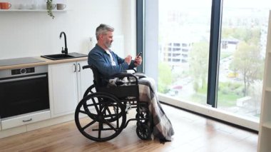 Tekerlekli sandalyede oturan orta yaşlı bir adam büyük apartman pencerelerinin yanında akıllı telefon kullanıyor.