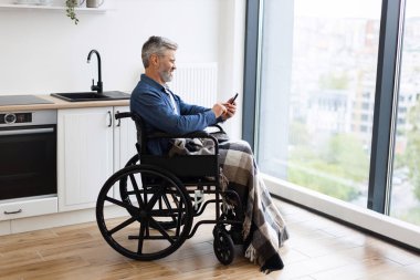 Tekerlekli sandalyede oturan orta yaşlı bir adam büyük apartman pencerelerinin yanında akıllı telefon kullanıyor.