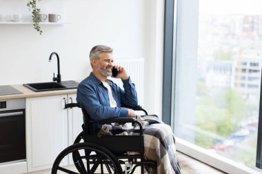 Üst düzey beyaz adam, pencerenin yanındaki telefonu kullanarak mutfağın yanında tekerlekli sandalyede oturuyor. Sahne günlük yaşam tarzı, erişilebilirlik, konfor, bağlantı, iletişim, bağımsızlık ve dahil edilmeyi betimler