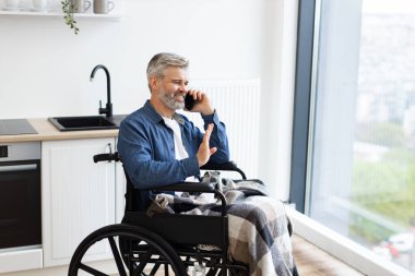 Üst düzey beyaz adam, pencerenin yanındaki telefonu kullanarak mutfağın yanında tekerlekli sandalyede oturuyor. Sahne günlük yaşam tarzı, erişilebilirlik, konfor, bağlantı, iletişim, bağımsızlık ve dahil edilmeyi betimler