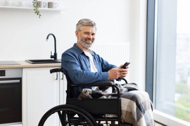 Parlak bir mutfakta akıllı telefon kullanarak tekerlekli sandalyede oturan olgun bir adam. Olumlu duygu, ulaşılabilirlik, bağımsızlık ve kıdemli engelliler için uygun modern yaşam tarzı gösteriyor