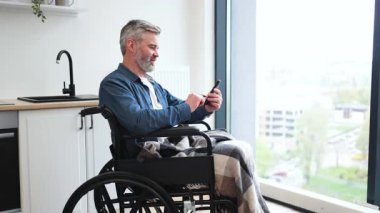 Üst düzey beyaz adam, pencerenin yanındaki telefonu kullanarak mutfağın yanında tekerlekli sandalyede oturuyor. Sahne günlük yaşam tarzı, erişilebilirlik, konfor, bağlantı, iletişim, bağımsızlık ve dahil edilmeyi betimler