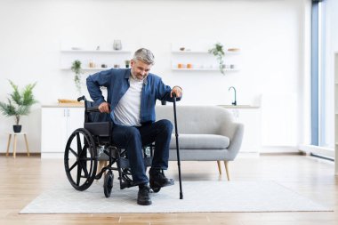 Tekerlekli sandalyenin yanında baston kullanarak yürüme pratiği yapan engelli bir adam. Ev ortamında bağımsızlık, kararlılık ve adaptif rehabilitasyon vurgulandı.