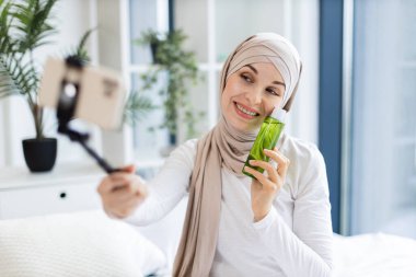 Genç Müslüman kadın blogcu, yüz nemlendiricisini destekleyen akıllı telefon özçekim çubuğuyla video kaydediyor. Doğal ışıklandırma ve yeşil bitkilerle donatılmış bir ev..