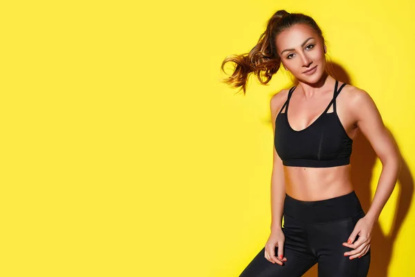 Young happy fitness girl with sporty body posing at studio on a yellow background. Beautiful fit Girl. Fitness smiling model in black sportswear. Weight Loss. Healthy lifestyle. Sporty healthy female. Horizontal