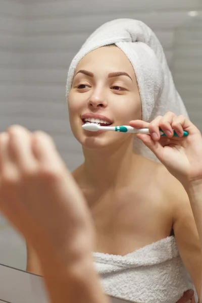 Young beautiful woman with towel on her head brushing her teeth at mirror