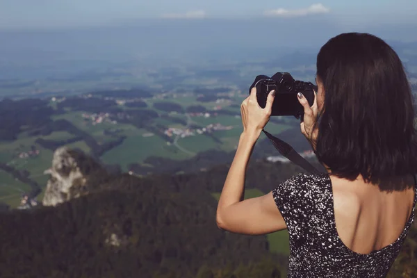 Brunette girl in a dress standing on the top of mountain and taking pictures with a camera.