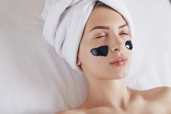 Young beautiful spa woman with towel on her head applying black mask cleaning pores. Skin care, spa salon and beauty Treatments.