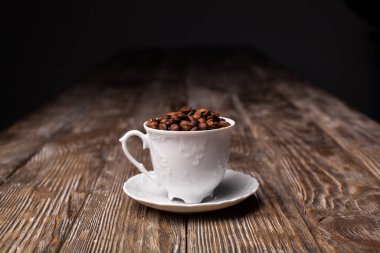 Beyaz bir kahve fincanı tam kahve çekirdekleri ile doludur. Fincanın üzerinde kahverengi desenli bir masa var.