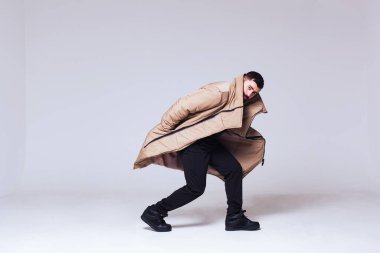 Beyaz arka planda bej renkli ceketli bir erkek model. Genç adam kışlık, sıcacık, açık renkli bir ceket giyiyordu. Yüzünün yarısını bir tasmanın arkasına sakladı. Model dinamik bir şekilde donup kaldı..