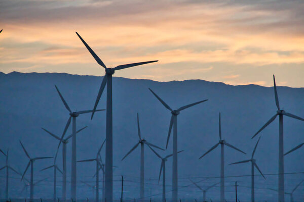 Green energy of wind power being harvested during the golden hour with a warm hued sky from a rising sun behind distant mountains.