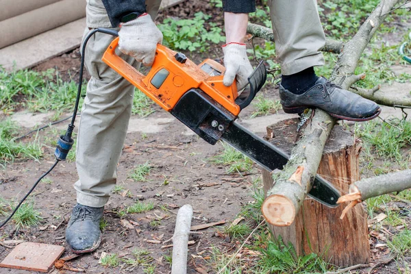 A man is sawing a log with an electric saw. Orange electro saw. Men\'s hands hold an electric saw, sawing a tree trunk.