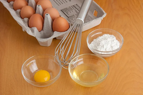 Separated cracked eggs in glass bowls, flour in a glass bowl, a silver whisk, and carton of eggs all on a cutting board.