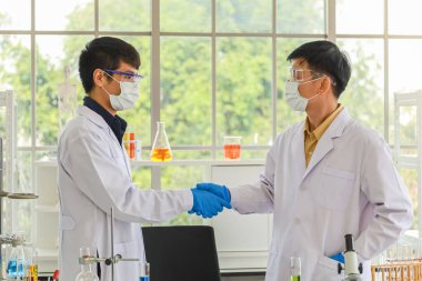 Tıbbi partikül koruyucu maske takan ve araştırma laboratuarında el sıkışan iki Asyalı uzman ve genç doktor..