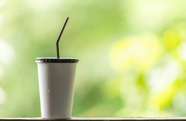 Paket kağıttan yapılmış, plastik kaplı buzlu kahve ve yeşil doğa arka planına sahip ahşap masa üzerinde saman ve kopyalama alanı..