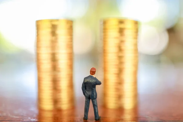 Business, Money Investment and Planning Concept.  Close up of businessman miniature people figure standing and looking to two stack of gold coins on wooden table with copy sapce.