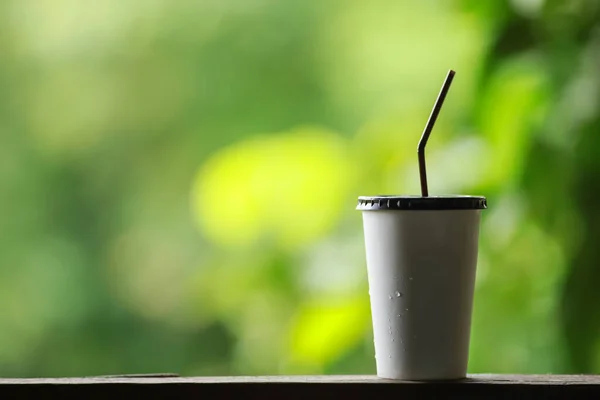 Closeup of takeaway paper cup of iced coffee with plastic cover and straw on wooden table with green nature background and copy space.