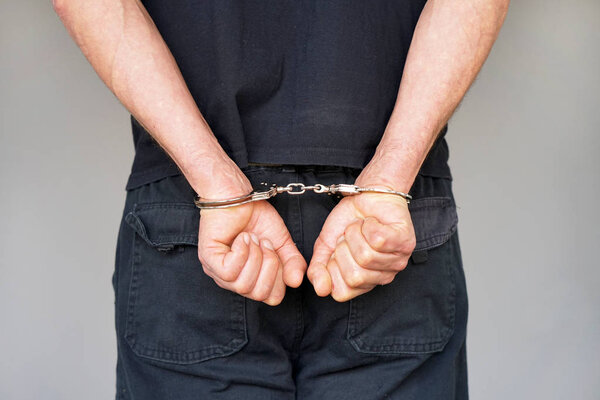 Close-up. Arrested man handcuffed hands at the back isolated on gray background. Prisoner or arrested terrorist, close-up of hands in handcuffs, selective focus.