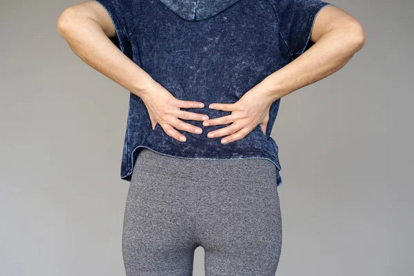 Close-up view of a young woman with pain in kidneys isolated on a gray background. Young woman with back ache clasping her hand to her lower back. Woman suffering from ribbing pain, waist pain.