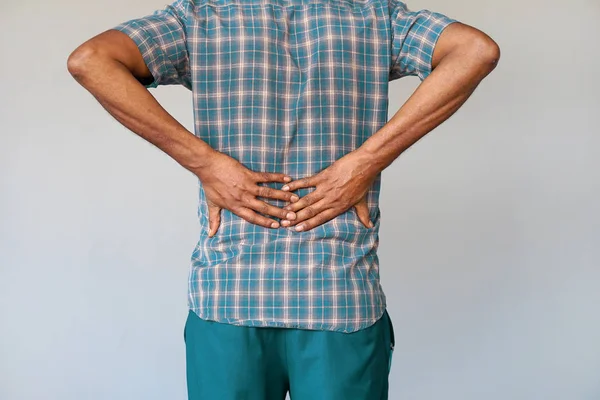 Close-up view of a young man with pain in kidneys on gray background. Young man with back ache clasping her hand to her lower back. Man suffering from ribbing pain, waist pain.