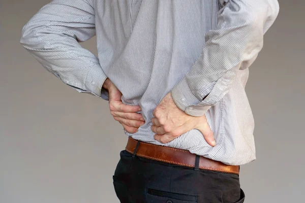Close-up view of a young man with pain in kidneys on gray background. Businessman with back ache clasping her hand to her lower back. Man suffering from ribbing pain, waist pain. Body Concept.