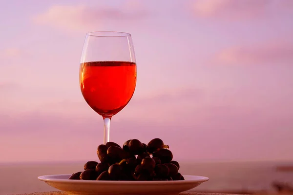 A glass of red wine standing on a plate of black grapes against a background of a sea horizon at sunset. A glass of red wine against the background of the evening sea horizon at sunset.