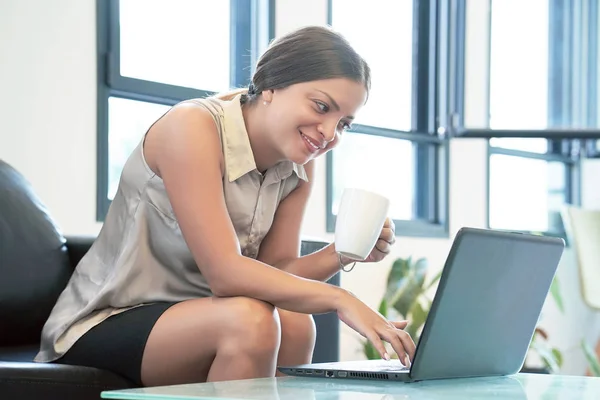 A woman sitting on the couch, smiling as she uses her laptop and hold a white coffee cup in her hand. Woman use her laptop for working and drinking cup coffee in hotel room. Cozy business atmosphere.