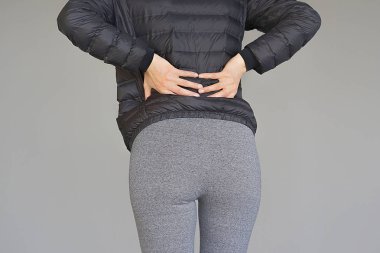 Gri bir arka plan üzerinde izole böbrek ağrısı olan yakın çekim kadın. Genç kadın elini sırtının alt clasping sırt ağrısı ile. Lastik ağrısı, bel ağrısı acı kadın.