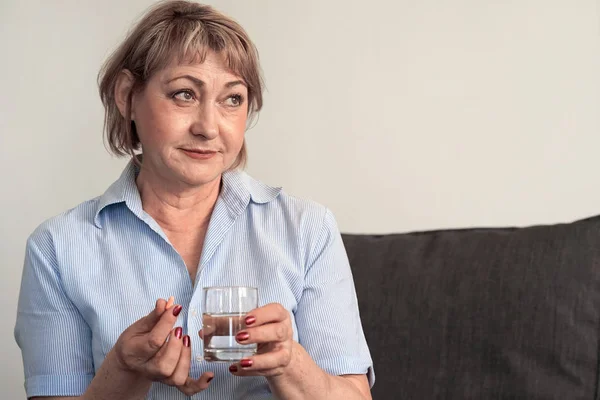生病的老年妇女拿着药丸杯的水在家里的沙发上 抑郁症不健康的妇女 即将采取抗抑郁药 紧急避孕 止痛药的痛苦时期 — 图库照片