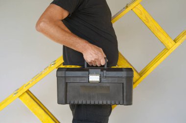 Elektrikçi araç kutusu ve sarı ahşap merdiven tutar. Builder bir ahşap merdiven ve araç kutusu gri bir arka plan üzerinde taşır. Araç kutusu ve sarı ahşap merdiven ile doğurmak.