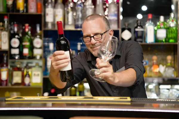 The bartender holds a bottle of red wine and wine glasses in the hotel bar. A bartender man gives a waiter a bottle of wine and glasses. The service concept.