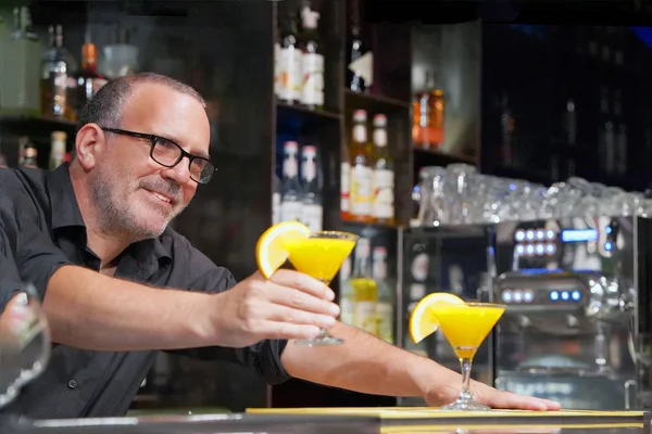The barman gives a cocktail to the client of the hotel bar. Fresh yellow cocktail with orange. Alcoholic, non-alcoholic drink-beverage at the bar counter in the night club. Glass of orange cocktail.