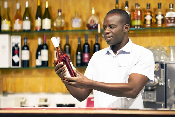 African bartender is holding a bottle of red wine. The barman examines a bottle of red wine.The bartender pours red wine to the client in the hotel bar. The concept of service. Concentrate on the bartender.